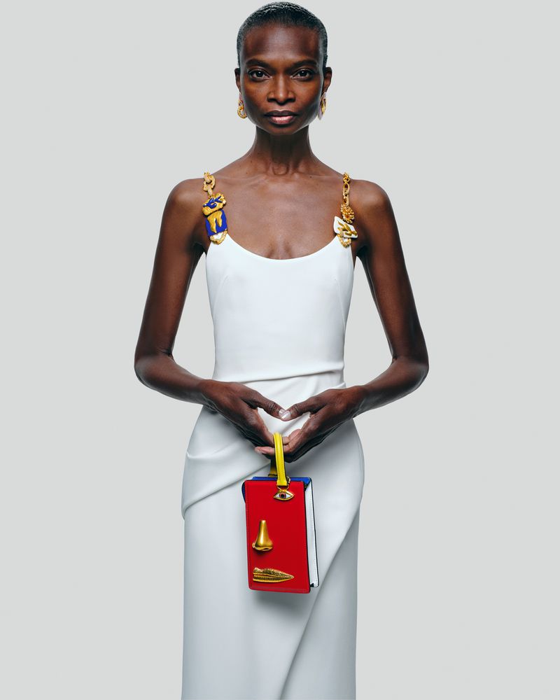 A slender Black female model poses in a white dress, holding a red purse with yellow handle against a grey backdrop in a studio shot.