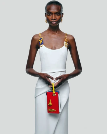 A slender Black female model poses in a white dress, holding a red purse with yellow handle against a grey backdrop in a studio shot.