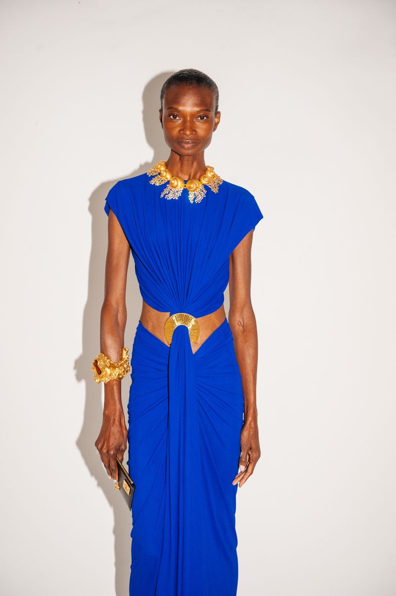 Elegant African woman poses in a vibrant blue gown and gold jewelry against a white backdrop in a fashion forward editorial shot.