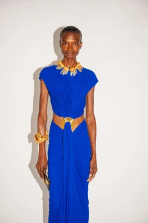Elegant African woman poses in a vibrant blue gown and gold jewelry against a white backdrop in a fashion forward editorial shot.