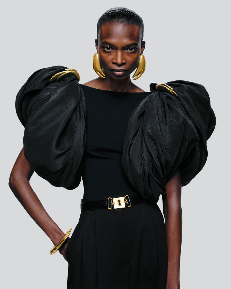 A striking portrait of a Black woman in a black top with dramatic puffed sleeves and large gold earrings, against a light gray backdrop.