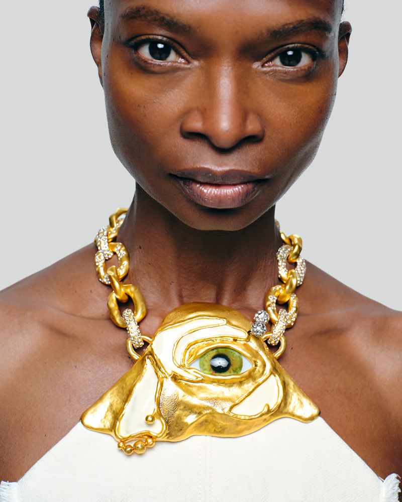 Close-up of a Black woman showcasing a distinctive gold statement necklace with a central eye design against a neutral background.