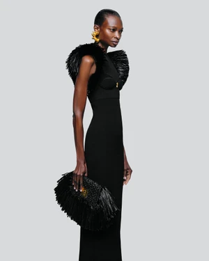 Model Debra Shaw wearing a black evening dress with textured shoulder details and gold earrings, holding a fringe clutch against a grey seamless background for Schiaparelli