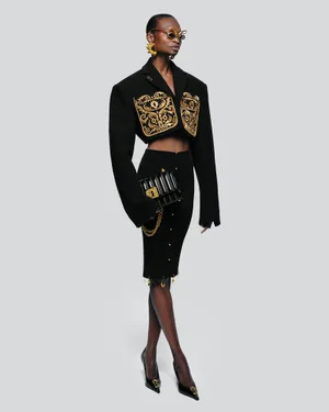 Model Debra Shaw wearing a black cropped jacket with gold embroidery and a pencil skirt against a seamless grey backdrop