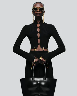 Model wearing gold sunglasses, statement earrings, and a black cutout top holding a large structured black handbag.