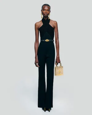 Model Debra Shaw wearing a black halter jumpsuit and holding a gold handbag against a plain grey backdrop.