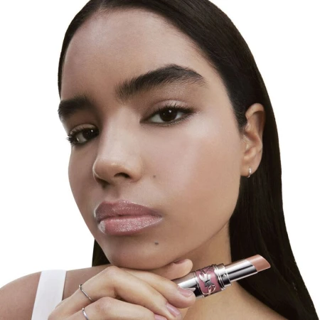 A woman holds a nude lip product, displaying glossy lips, fair skin, and a natural makeup look in a beauty or cosmetic advertisement.