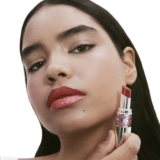 Adult woman posing indoors holding a YSL lipstick tube close up. The model wears glossy lipstick in a beauty shot against white.