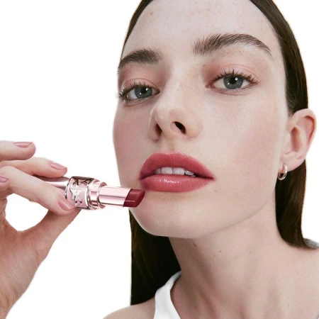 Close-up of a woman applying berry-toned lipstick to her lips, showcasing makeup application in a beauty context on a white background.