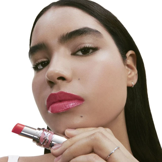 Close-up of a young woman with pink lipstick holding a YSL lipstick tube in a beauty advertisement against a white background.