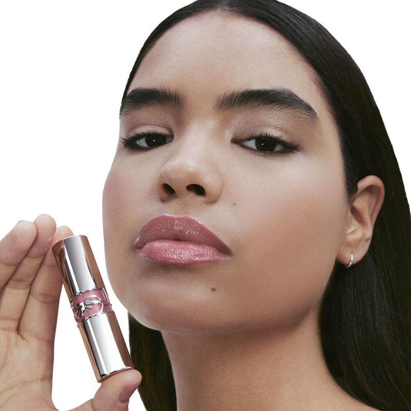 Close-up of a woman with dark hair holding a shiny tube of lip gloss, showcasing glossy lips and radiant skin in a beauty product shot.