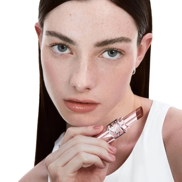 Close-up of a young adult female holding a tinted lip balm, showcasing its color and packaging against a white background.