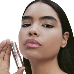 Close-up beauty portrait of a woman holding a metallic YSL Loveshine lipstick tube near her lips against a solid white background.