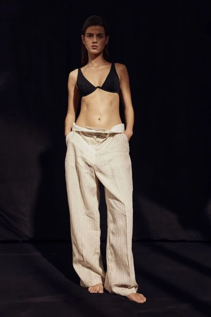 Adult woman poses in a black bikini top and loose-fitting striped pants against a dark background in a fashion photography studio.