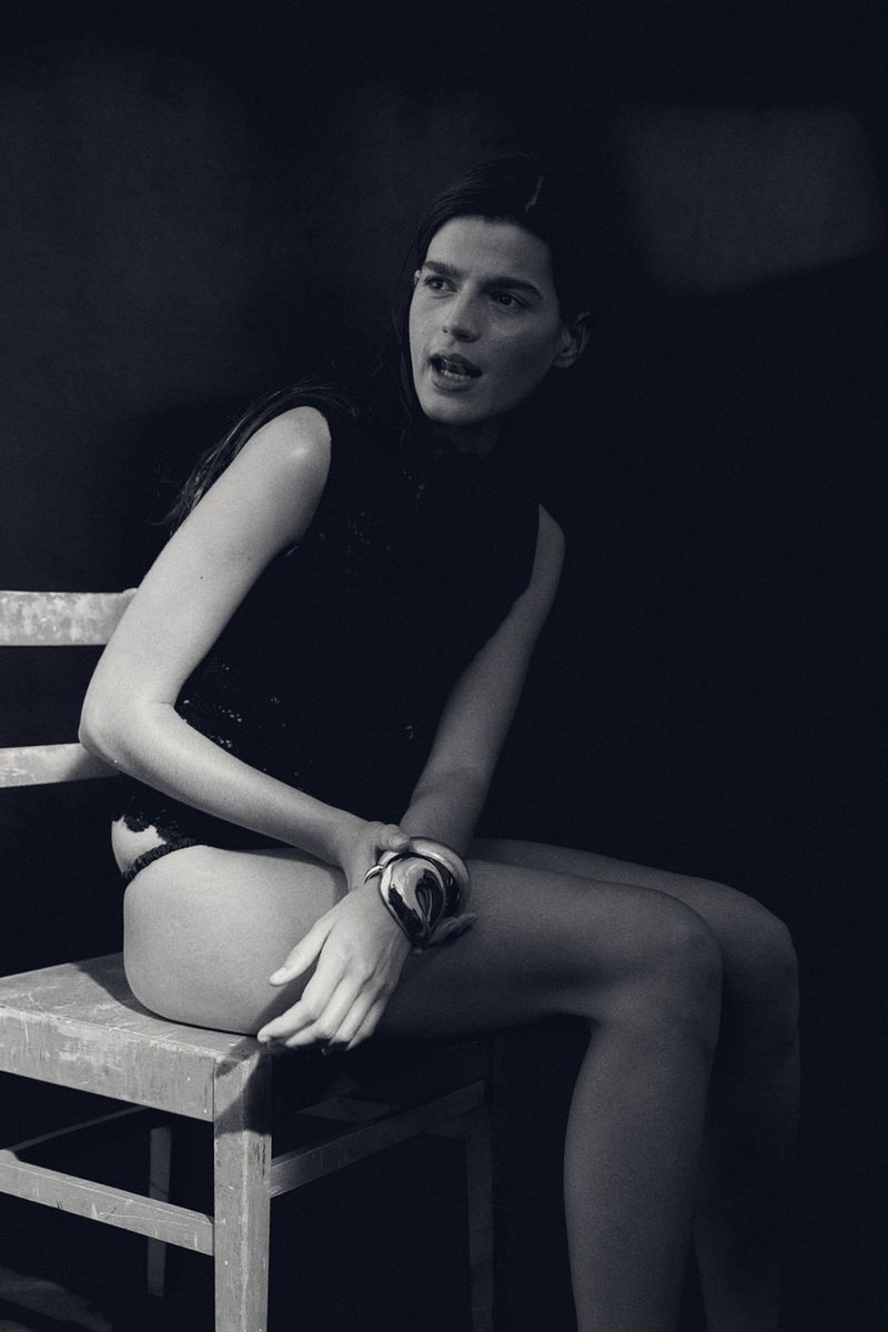 Monochrome portrait of an adult female fashion model sitting on a wooden chair in a dark photo studio, wearing a top and bracelet.