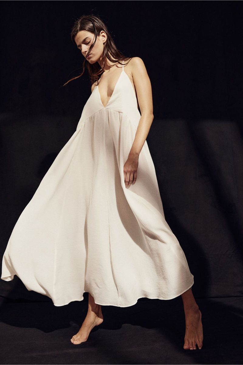An adult female model wears a white dress in a studio. She is barefoot. The dress is flowy with spaghetti straps against a dark background.