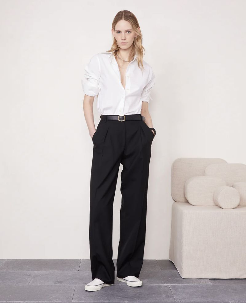 Full-length shot of a fair-skinned woman in a white shirt and black trousers, posing in a neutral indoor setting, fashion style.