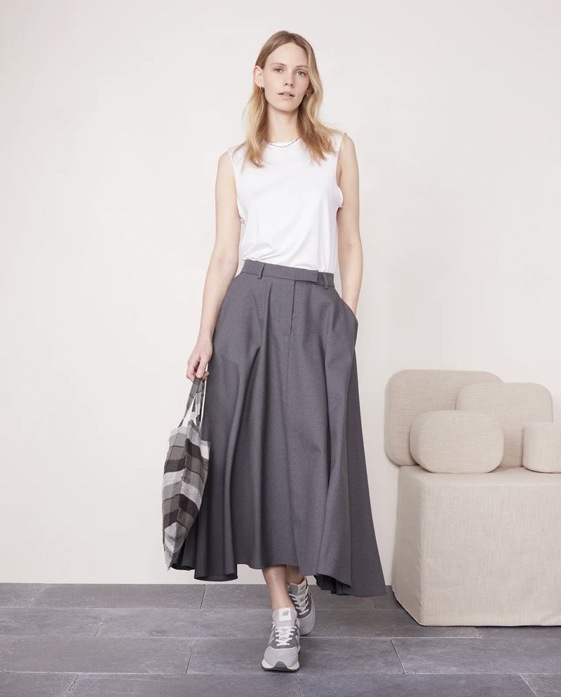 Full shot of a blond female model in a white sleeveless top and grey pleated skirt holding a tote bag and wearing trainers.