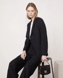 Model seated on a wooden stool wearing a black suit jacket and trousers holding a black handbag against a light studio backdrop.