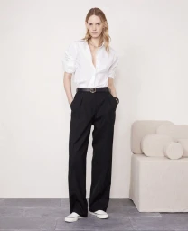 Model wearing a white button-down shirt and black trousers standing against a plain light wall with a modern beige stool.