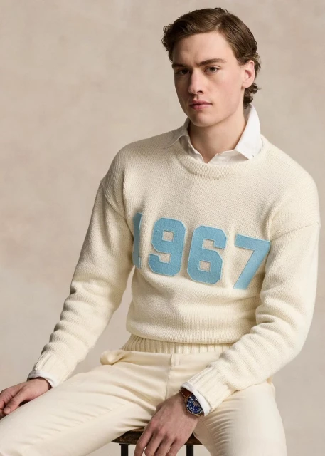 A fair-skinned young man poses indoors, wearing an off-white sweater with '1967' in blue numerals, cream pants, and a wristwatch.