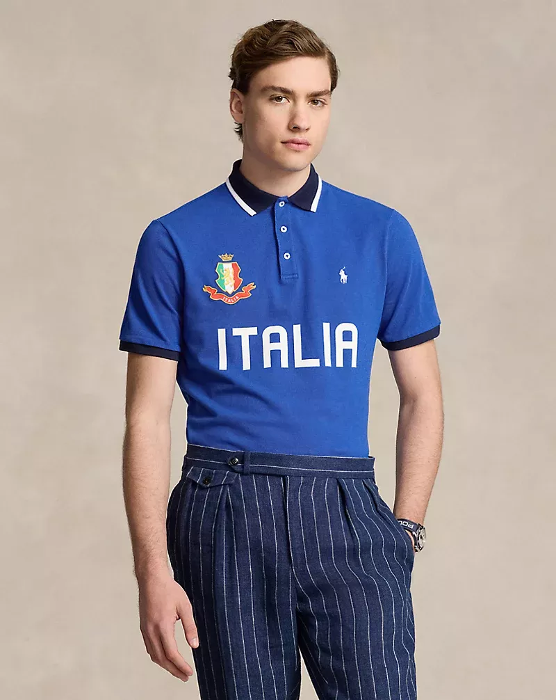 A fair-skinned male model wears a blue polo shirt with an Italian crest and white pinstripe dress pants, posing in a studio setting.