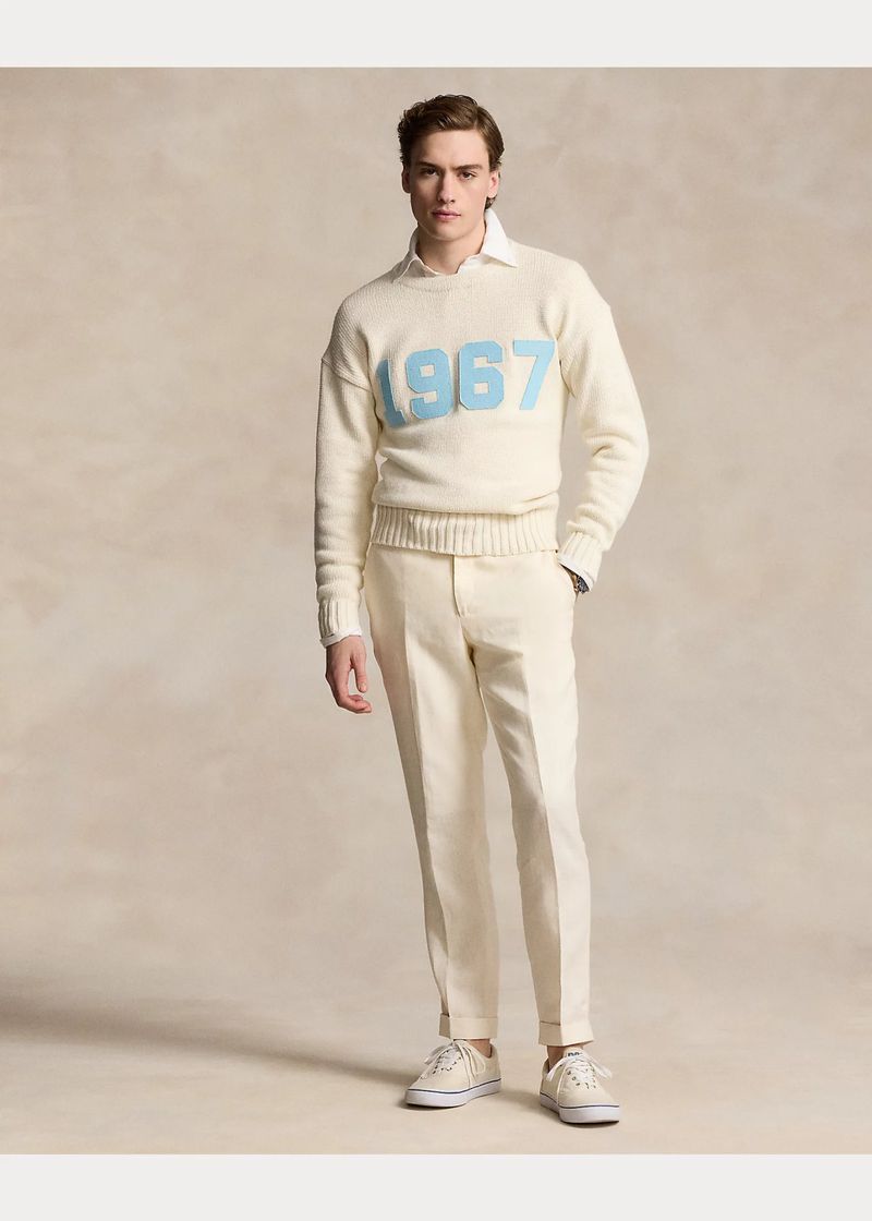 Full shot of a male model in a cream-colored sweater with '1967' in light blue, cream-colored pants and sneakers on a neutral background.