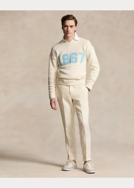 Full shot of a male model in a cream-colored sweater with '1967' in light blue, cream-colored pants and sneakers on a neutral background.