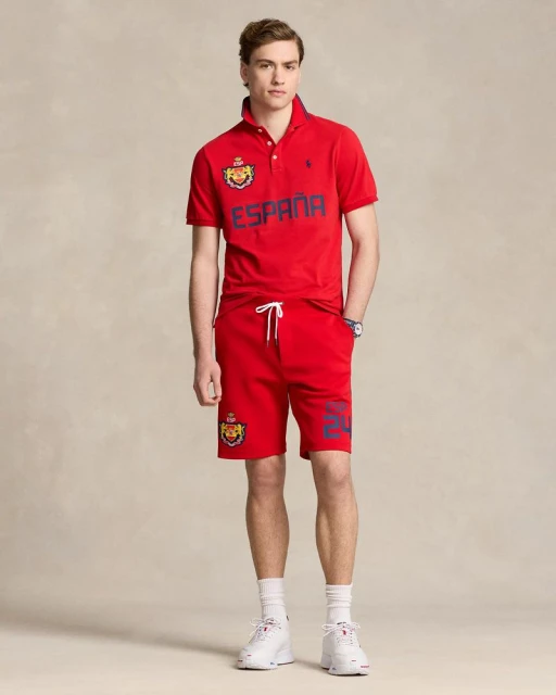Man in red Ralph Lauren polo shirt and shorts with ESPAÑA text and crests. He wears white sneakers and socks in a studio shot.