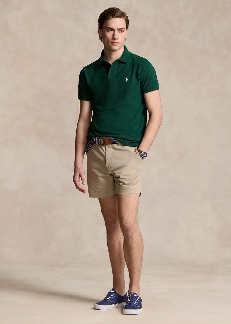 Full-length shot of an adult male wearing a green polo shirt with khaki shorts and blue sneakers, posing against a neutral backdrop.