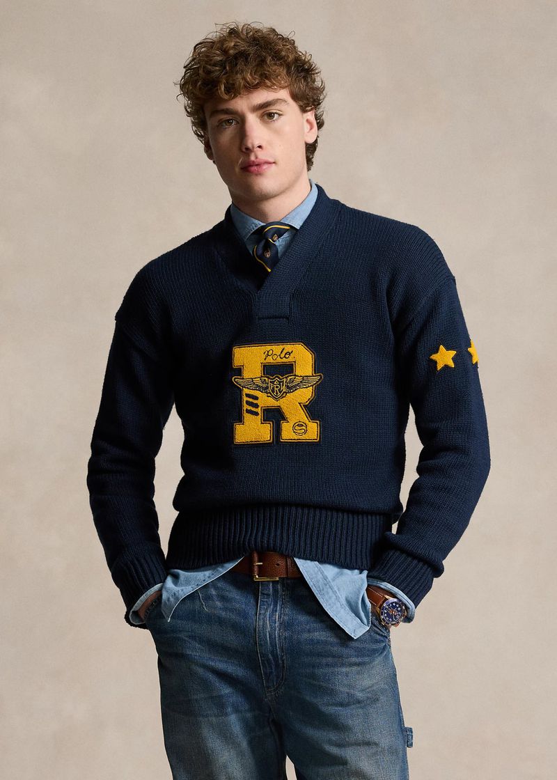 A young adult male poses indoors wearing a navy blue knit sweater with a graphic logo and blue jeans in a waist-up portrait.