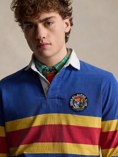 Adult male with curly brown hair wears a blue, yellow, and red striped rugby shirt with a Polo S.R.L. Alpine Club patch.