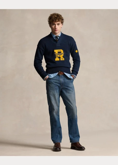 Front-facing studio shot of a young adult man wearing a navy V-neck sweater with a light blue collared shirt and dark-wash jeans.