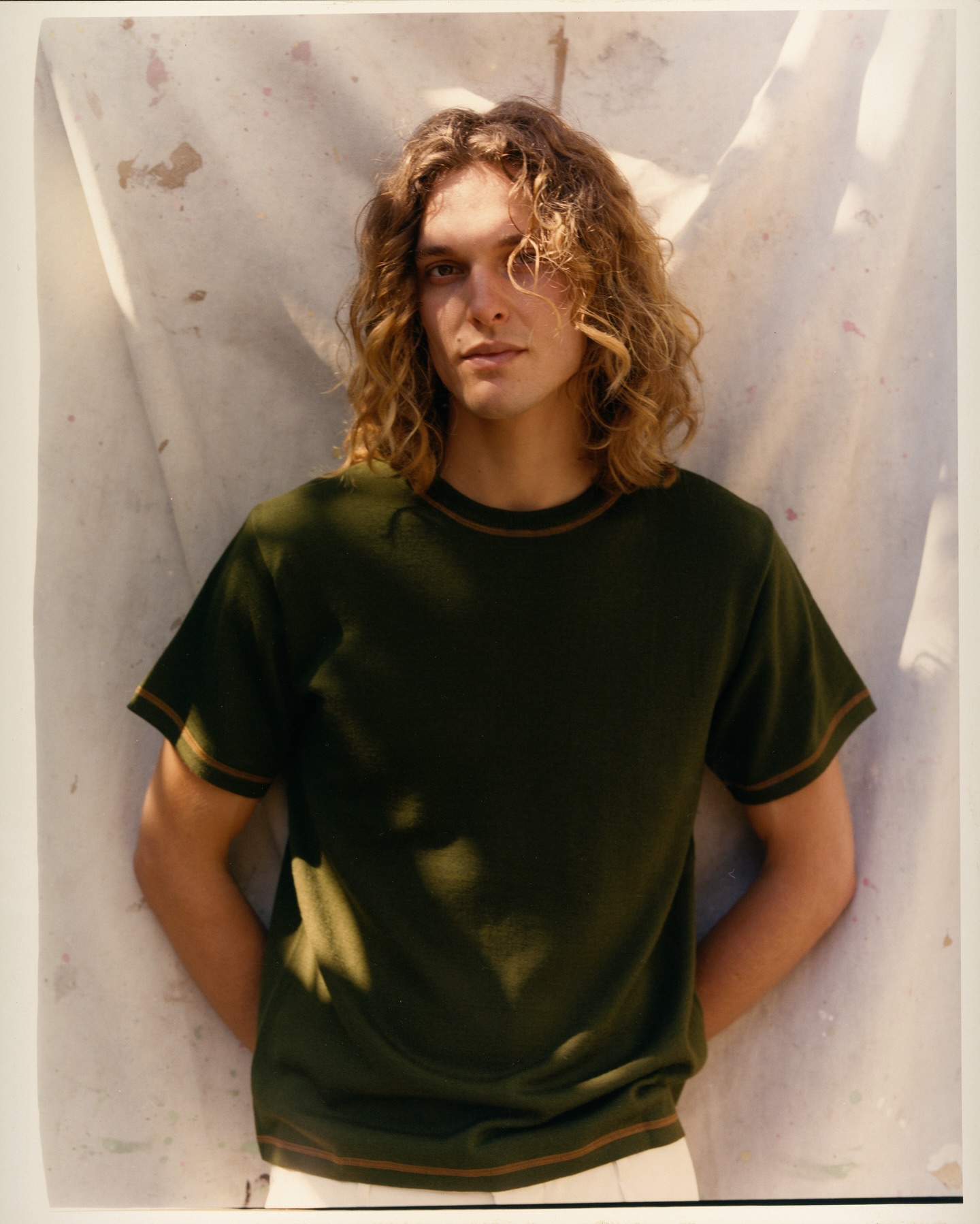 Indoor portrait of a young adult male with wavy blond hair, wearing a dark green t-shirt against a textured off-white backdrop.