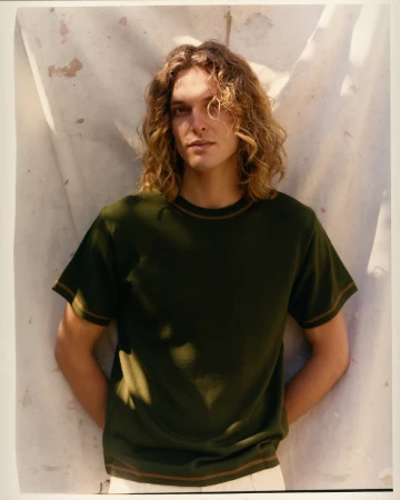 Indoor portrait of a young adult male with wavy blond hair, wearing a dark green t-shirt against a textured off-white backdrop.