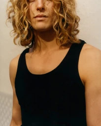 Portrait of a man with curly blonde hair wearing a black tank top against a neutral background.
