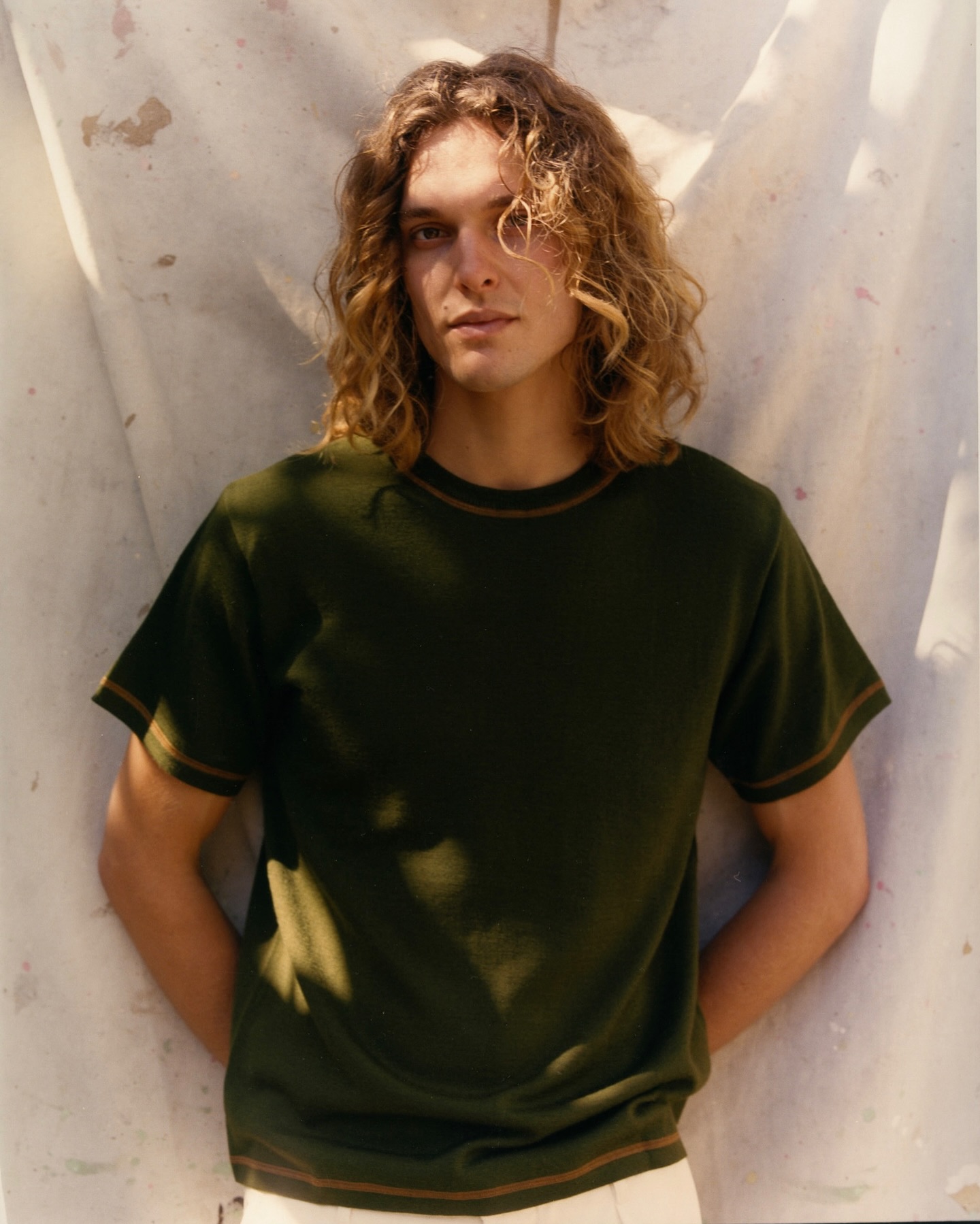 Portrait of a young adult male with long, wavy hair wearing a dark green T-shirt in front of a textured off-white backdrop.