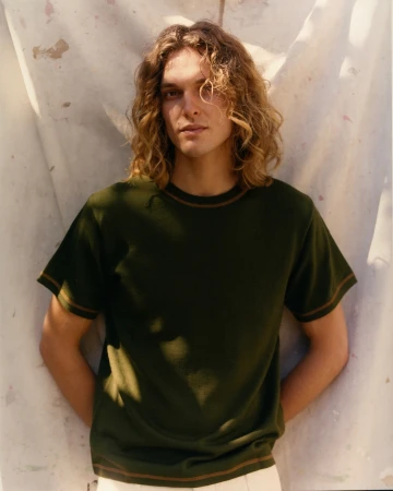 Portrait of a young adult male with long, wavy hair wearing a dark green T-shirt in front of a textured off-white backdrop.