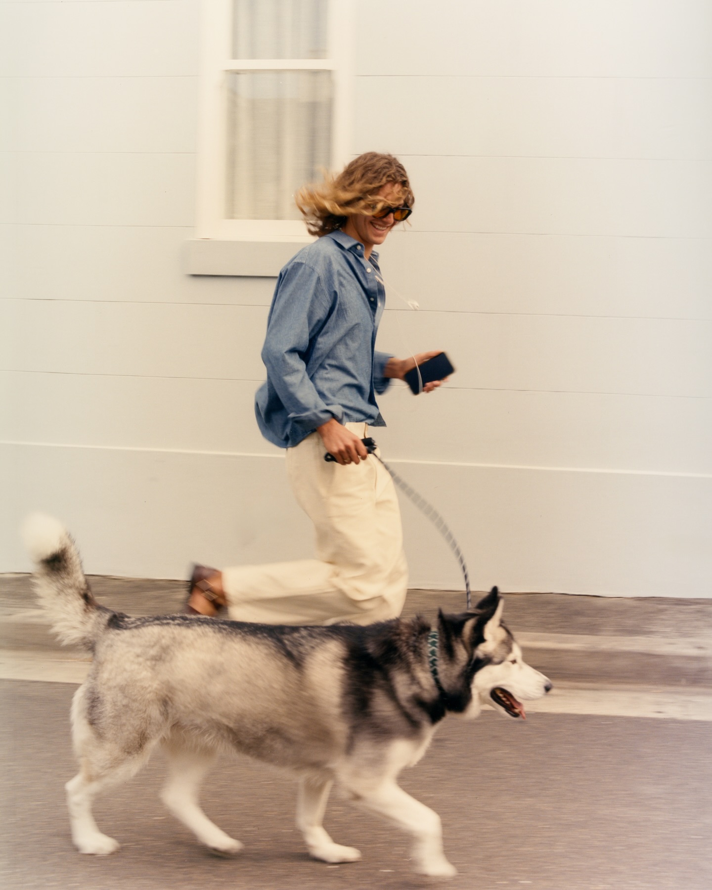 Woman jogging with a grey and white Siberian Husky on a leash on a sidewalk near a white building with a window.