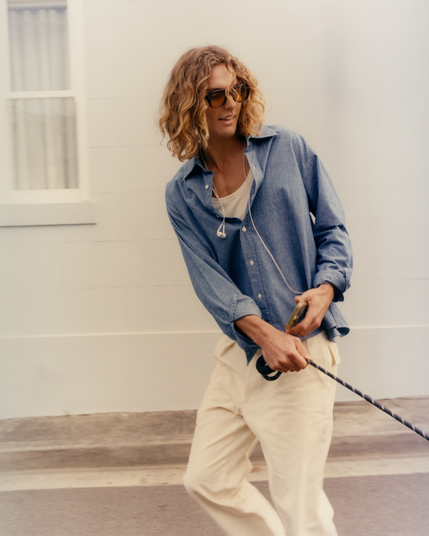 An adult male with long blonde hair and sunglasses in a blue shirt and white pants holds a black leash, walking outside in daylight.