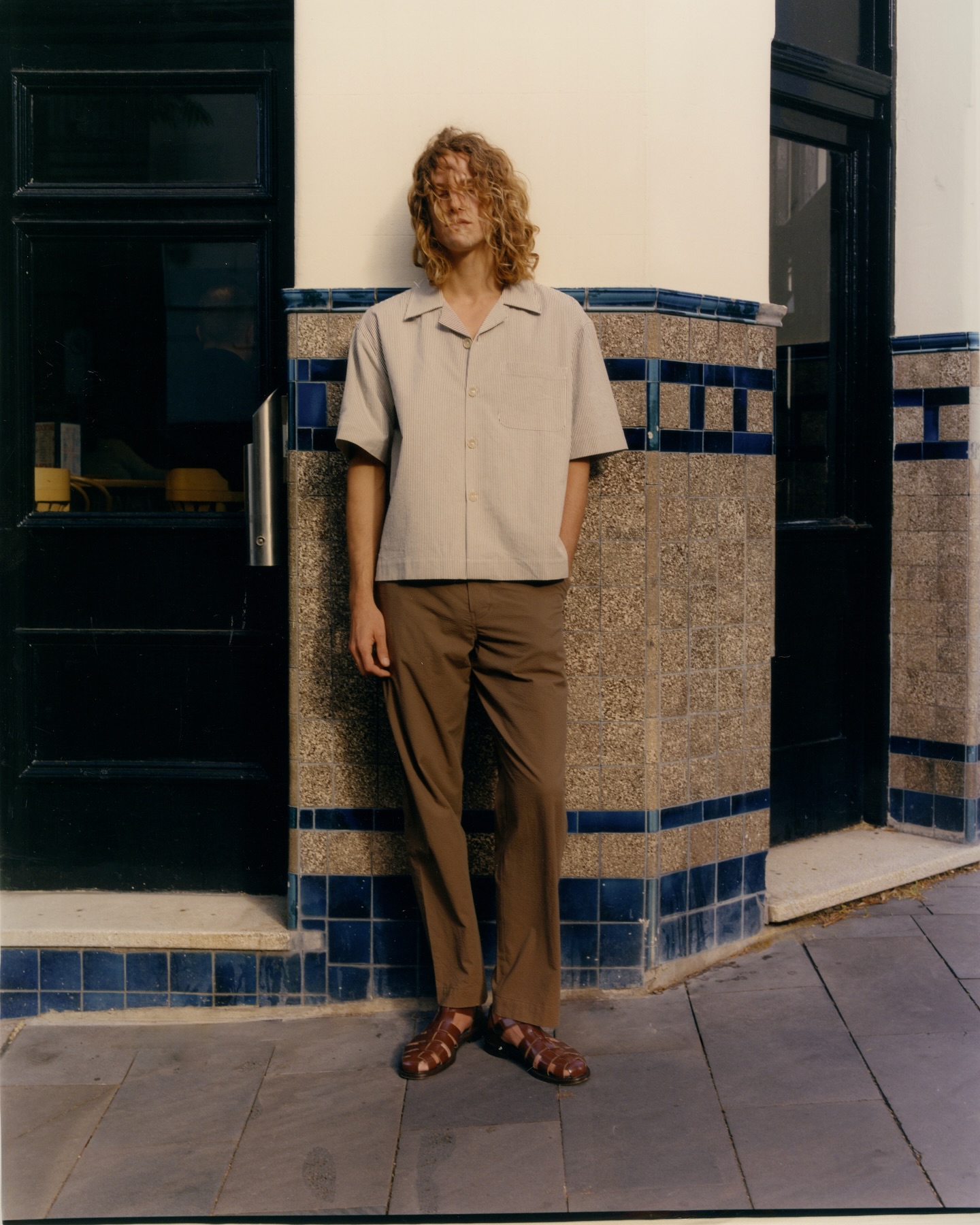 Fashion shot of a male model in a beige short-sleeved shirt and brown pants leaning against a blue and white tiled wall outdoors.