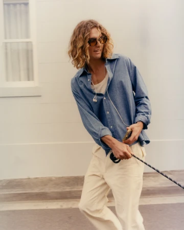 A young adult male with long curly blond hair walks outside, wearing an open blue shirt, white pants and sunglasses while holding a leash.