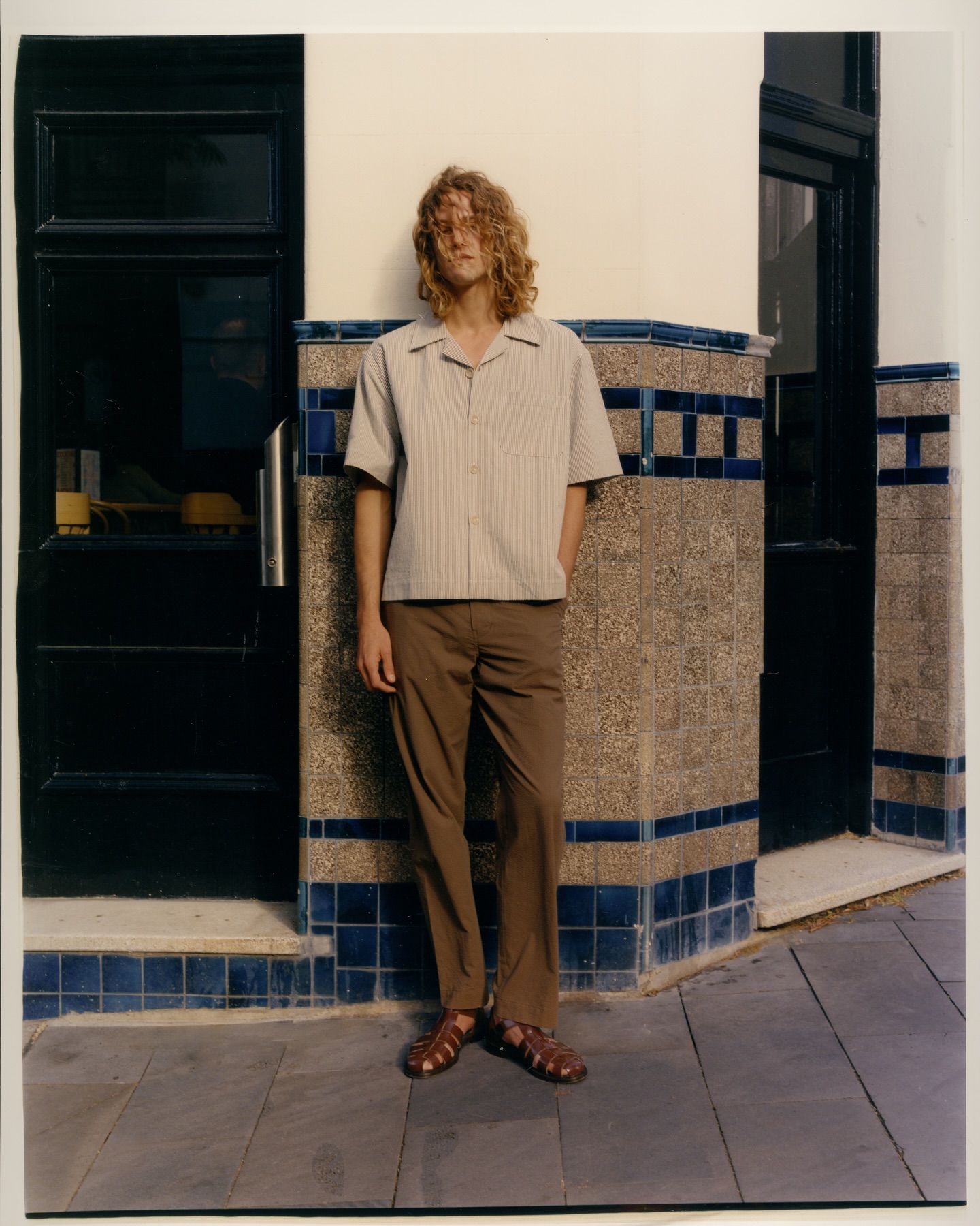 Full shot of a man with curly hair wearing a button-down shirt, brown pants, and sandals, standing against a tiled wall outdoors.