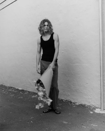 Black and white portrait of a man with long curly hair wearing a dark tank top holding a wrapped bouquet against a light wall