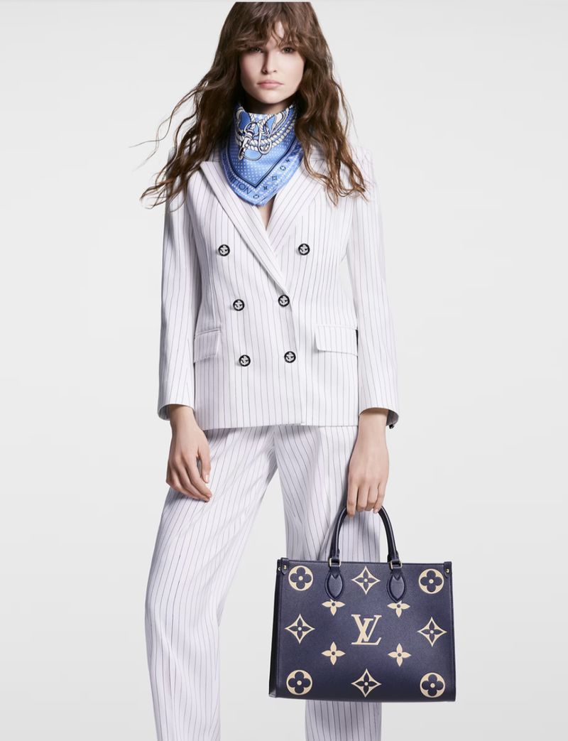 A woman in a pinstripe suit holds a Louis Vuitton handbag against a white background in a fashion shot with soft studio lighting.