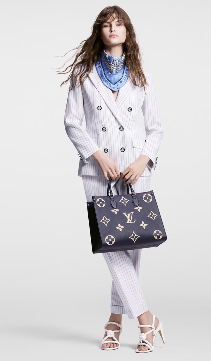 A full shot of a woman in a white pin-striped suit holding a dark blue Louis Vuitton handbag against a bright white backdrop.