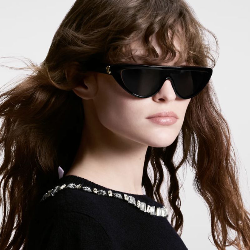 A woman with long brown hair wears black sunglasses and a dark sweater with jewel embellishments against a neutral backdrop.