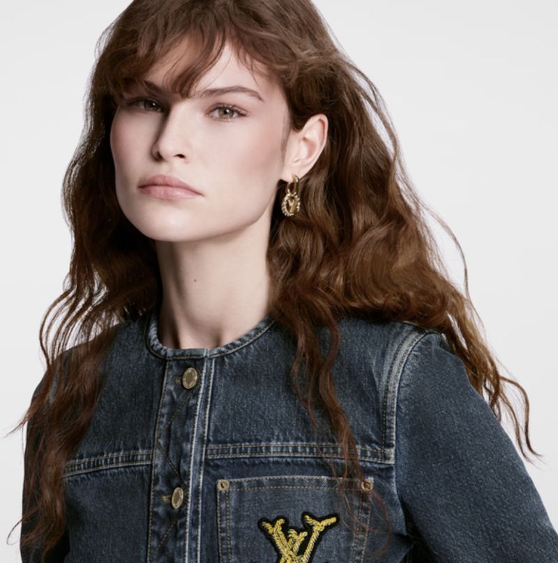 A woman with long, wavy brown hair wears gold earrings and a blue denim jacket with a logo patch on the pocket against a white background.