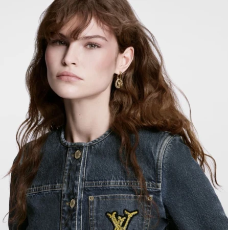 A woman with long, wavy brown hair wears gold earrings and a blue denim jacket with a logo patch on the pocket against a white background.