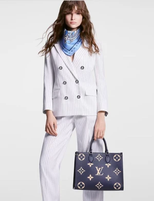 Model wearing a white pinstripe suit and blue silk scarf holding a dark blue patterned monogram handbag against a white background.